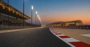 Empty floodlit race circuit at dusk with red and white kerbs and a lattice-roof hotel in the distance